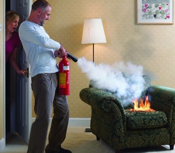 Person putting out a couch fire with a fire extinguisher in their home while another person watches in shock.